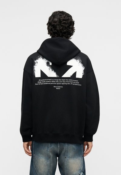 Άτομο με κατσαρά μαλλιά φοράει μαύρο φούτερ με κουκούλα με λευκά αφηρημένα βέλη και τυπωμένο κείμενο στην πλάτη, συνδυασμένο με ξεθωριασμένο μπλε τζιν.