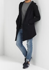 Longue veste à capuche noire, avec une coupe ajustée, portée sur une chemise grise. Associée à un jean bleu clair et des chaussures de sport noires.