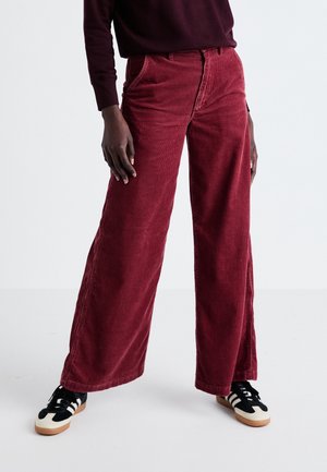 Person wearing high-waisted wide-leg red corduroy pants, dark long-sleeve top, and black-and-white sneakers, standing with legs apart.