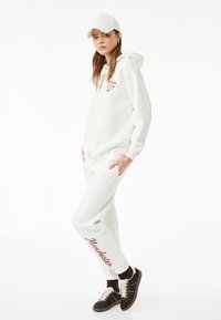 Sudadera blanca con un logo en el frente, combinada con pantalones de chándal a juego que llevan el texto "Manchester" en marrón. Zapatillas marrones completan el conjunto.