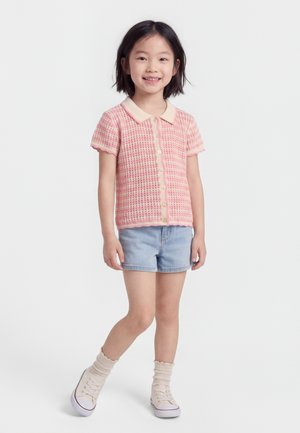 Mladé dievča sa usmieva, nosí ružovú krátku blúzku na gombíky, svetlomodré džínsové šortky, béžové ponožky a biele tenisky, stojí pred bielym pozadím.