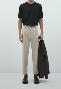 Svart kortärmad t-shirt, beige skräddarsydda byxor med sidofickor, svarta läderskor och en mörk jacka som hålls i höger hand.