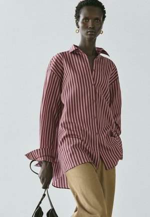 Vrouw draagt een oversized roze en beige shirt met verticale strepen, beige broek, gouden oorringen en houdt een zwarte handtas vast tegen een grijze achtergrond.