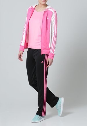 Femme portant un sweat à capuche rose, un t-shirt rose assorti, un pantalon de survêtement noir avec des rayures roses, et des baskets bleu clair, debout devant un fond gris.