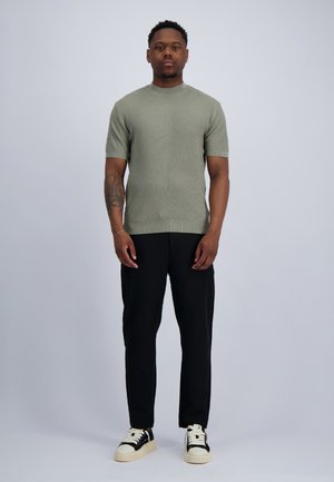 Man staat recht en kijkt naar voren, draagt een geribd groen shirt met korte mouwen, zwarte broek en zwart-witte sneakers tegen een effen grijze achtergrond.