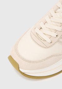 Beige tossude ja sissetõmmatava mesh-kanga tekstuuriga sneiteri nina lähedalt, kreemivärvi paelad ja kummine tall valgel taustal.