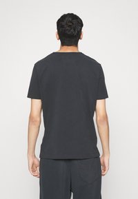 Homme portant un t-shirt noir uni à manches courtes et un pantalon noir, se tenant de dos face à l'appareil photo sur un fond blanc.