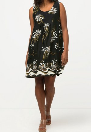 Kobieta ubrana w czarną, bezrękawnikową sukienkę z białymi i złotymi kwiatowymi oraz łańcuchowymi wzorami, do której dobrano sandały na koturnie z przezroczystymi paskami.