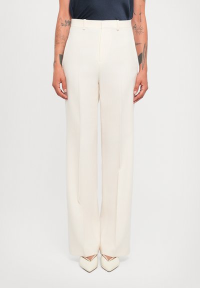 Pantaloni bianchi su misura con vita alta e gamba larga, caratterizzati da una texture liscia, pieghe sottili e una finitura pulita. Abbinati a tacchi color crema.