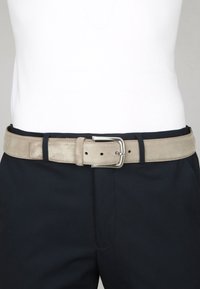 Beige suède riem met een rechthoekige zilveren gesp, voorzien van verschillende gaatjes voor verstelbare pasvorm, gedragen met donkere broek.