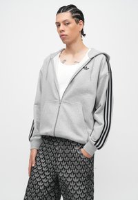 Pilkas gobtuvu užsegamas džemperis su užtrauktuku, juodomis juostomis ant rankovių ir mažu logotipu krūtinės srityje, derinamas su juodomis raštuotomis kelnėmis.