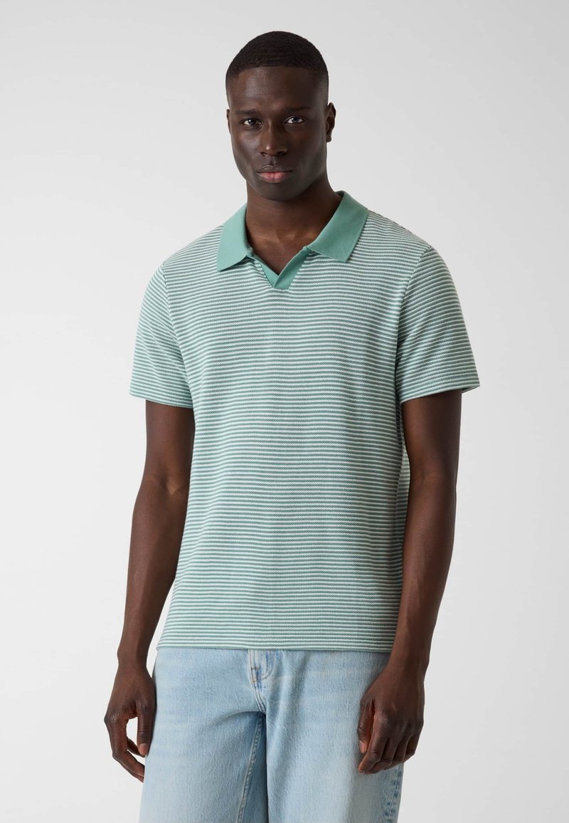 Homme debout, portant un polo vert clair à rayures avec un col vert uni et un jean bleu clair, face à un fond uni.