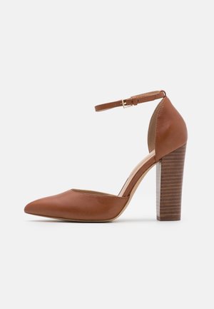 Brązowy skórzany najwyższy obcas z zakrytymi palcami, z paskiem na kostkę i klamrą, posiadający gruby, warstwowy drewniany obcas na białym tle.