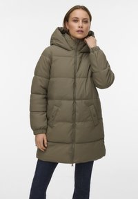 Kvinne iført en knelengde olivengrønn polstret kåpe med hette og glidelåslukking, kombinert med mørkeblå jeans, mot en ensfarget hvit bakgrunn.