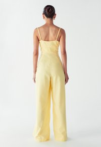 Mujer con el cabello recogido en un moño, vistiendo un mono sin mangas de pierna ancha en un tono amarillo pálido, de pie de espaldas contra un fondo claro y liso.