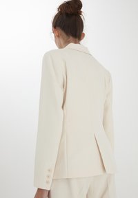 Beige skräddarsydd kavaj med en slits bak, långa ärmar och fem krämfärgade knappar vid ärmslutet. Slät tyg med en strukturerad design.
