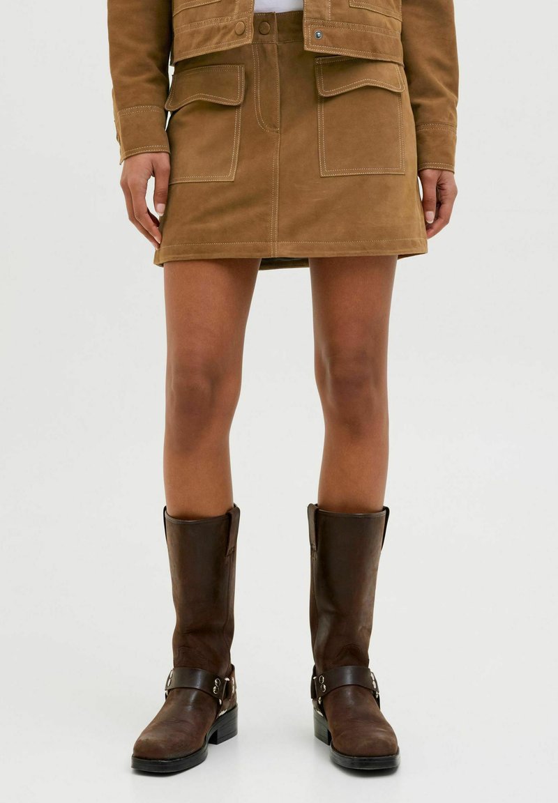Jupe mini en daim marron avec deux poches avant, complétée par des bottes en cuir marron foncé jusqu'au genou avec des boucles. Design simple et texture lisse.