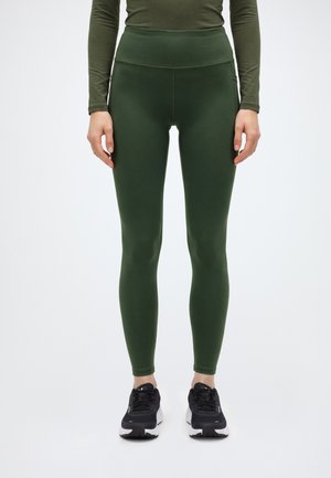 Un mannequin se tient sur un fond blanc, vêtu d’un legging vert foncé taille haute, d’un haut assorti à manches longues et de baskets noires.