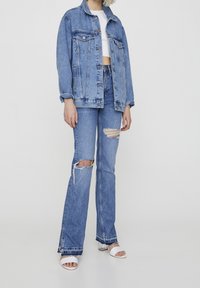 Lichtblauwe spijkerjas met een losse pasvorm, gecombineerd met lichtblauwe flared jeans met slijtageplekken. Witte sandalen met een blokhak maken de look compleet.