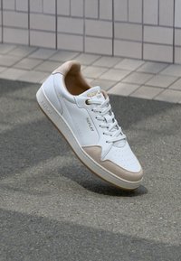 Weißes Ledersneaker mit beigen Wildlederakzenten. Merkmale sind eine perforierte Zehenpartie, flache Schnürsenkel und eine strukturierte Gummisohle. Markenlogo ist abgebildet.