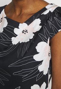 Primer plano de un vestido floral oscuro con grandes flores blancas y rosa pálido, y delgadas líneas de hojas blancas en el hombro y cuello de una persona.