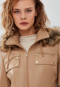 Tan faux leather jacket with a fur-lined hood, gold hardware, zippered chest pockets, and a textured finish.