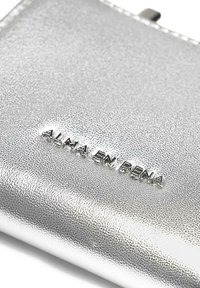 Cartera plateada con acabado texturizado, con el nombre de la marca en relieve "ALMA EN PENA" y un diseño elegante y minimalista.