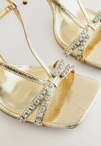 Zelta atvērtu purngalu augstpapēžu kurpes ar plānām metāliskām plaisām, kurām ir caurspīdīga rhinestone akcenta detaļa uz siksnām un kvadrātveida purngala forma.