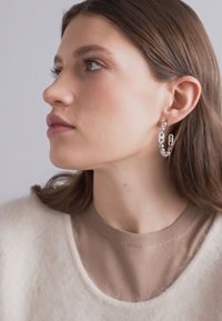Orecchini a cerchio in argento con anelli intrecciati, indossati da una persona con lunghi capelli castani. Sono abbinati a un maglione beige.