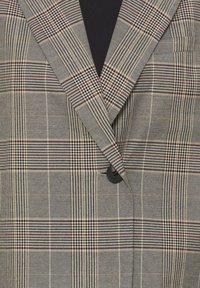 Gros plan d'un blazer gris à carreaux avec des lignes beige, noires et rouges, un seul bouton noir fermé à l'avant.