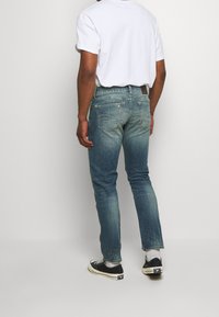 Man som bär urblekta blåjeans, vit t-shirt, vita strumpor och svarta sneakers, står mot en enkel ljus bakgrund vänd bort.