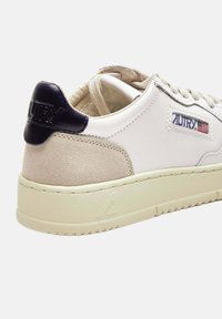 Scarpa da ginnastica bassa bianca e beige con toppa sul tallone blu navy, suola color crema, toppa con logo Autry e lacci parzialmente slacciati su sfondo bianco.