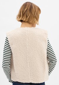 Nepbont vest in lichtbeige, met een zachte textuur. De achterkant heeft een naaddetail. Ggedragen over een gestreepte longsleeve.