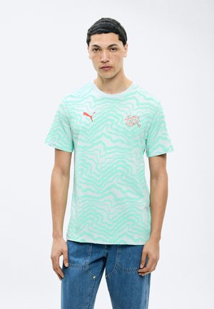 Νεαρός άνδρας που φοράει ανοιχτό πράσινο αθλητικό μπλουζάκι Puma με κόκκινο λογότυπο και μπλε τζιν, στέκεται μπροστά σε λευκό φόντο.