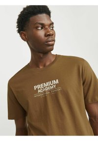 Brun bomulds t-shirt med et hvidt grafisk print, der indeholder teksten "PREMIUM ACADEMY" og "INTERNATIONAL CONCEPT". Korteærmer, rund hals design.