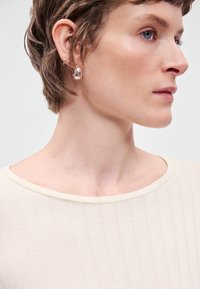 Közeli kép egy személyről, aki világos bézs bordázott felsőt visel, széles nyakkivágással, sima textúrával és finom bőrszínnel. Ezüst fülbevalók láthatók.