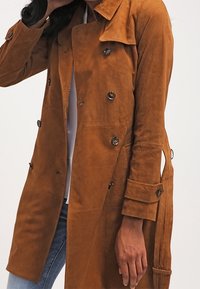 Brązowy zamszowy płaszcz trench z podwójnym zapięciem, dużymi guzikami i bocznymi kieszeniami, o dopasowanym kroju i uchwytach na pasek.
