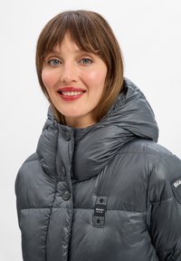 Doudoune grise avec une grande capuche rembourrée, des boutons-pression et un écusson "Blauer U.S.A." sur la manche. Texture lisse et brillante.