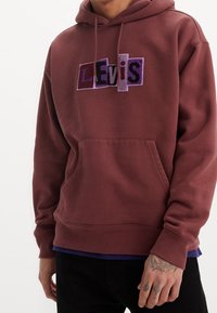 Muž v vínovém mikinovém svršku s fialovým nápisem "Levi's" na hrudi a viditelnými tetováními na pravé ruce na bílém pozadí.