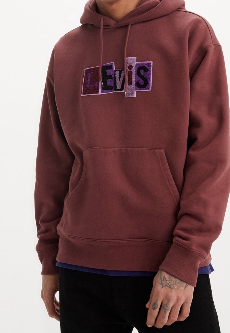 Muž v vínovém mikinovém svršku s fialovým nápisem "Levi's" na hrudi a viditelnými tetováními na pravé ruce na bílém pozadí.