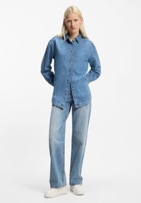 Camisa de mezclilla azul claro con cierre de botones al frente, mangas largas y cuello puntiagudo, combinada con jeans de talle alto azul claro y zapatillas blancas gruesas.