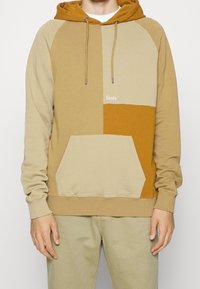 Sweat à capuche à blocs de couleur beige et marron en tissu doux, avec une poche kangourou, une capuche à cordon de serrage et un logo brodé sur la poitrine.