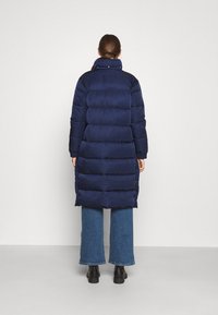 Mørkeblå dunjakke med høj krave og kviltet design, med en knælængde kant og sidelommer, båret over brede denimbukser.