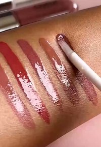 Vier glanzende lipgloss-stalen in naaktkleuren op de huid, waarbij een wit applicatorborsteltje de vierde swatch aanbrengt.