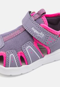 Lila und rosa Kindersandalen mit einem Obermaterial aus Mesh, verstellbarem Riemen, offener Zehenpartie und Gummisohle mit weißen Akzenten.