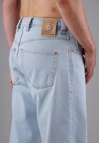 Hellblaue Denim-Jeans, die von einer Person getragen werden, mit sichtbaren Gesäßtaschen und einem Ledermarkenetikett in der Nähe des Bunds auf einem schlichten grauen Hintergrund.