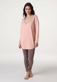 Tunika rosa chiaro a maniche lunghe con scollo a V e bordatura nera abbinata a leggings marroni aderenti. Texture liscia, design casual.