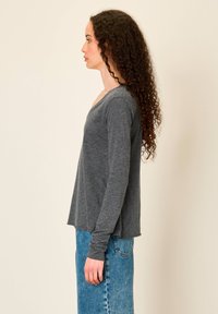 Moteris su ilgais garbanotais plaukais, dėvinčia pilką ilgomis rankovėmis megztinį ir mėlyną džinsinę sijoną, stovinti profilio pozicijoje priešais paprastą foną.