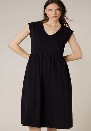 Femme aux longs cheveux bruns portant une robe noire midi sans manches avec taille froncée, debout les mains sur les hanches devant un fond beige uni.