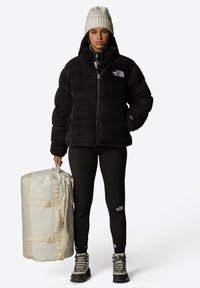 Czarna polarowa kurtka puchowa z zamkiem błyskawicznym i logo, zestawiona z czarnymi legginsami i szarymi butami turystycznymi. Kremowa torba typu duffel z ramiączkami.